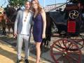 Una pareja elegante posa frente a un carruaje de caballos en un evento formal.