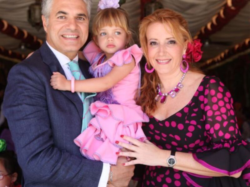 Una familia feliz en un ambiente festivo, con una madre y padre sonrientes sosteniendo a su hija pequeña vestida de rosa. La mamá lleva un vestido negro con lunares rosados y una pulsera de perlas, mientras que el papá está en un traje azul con corbata verde. La escena parece ser parte de una celebración o fiesta, con decoraciones en el fondo.