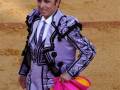 Un torero en traje tradicional de color púrpura y blanco, con un pañuelo rosa en la mano izquierda.