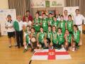 Equipo femenino de baloncesto celebrando su victoria en un torneo local, con medallas y una bandera nacional en el fondo.