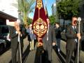 Un grupo de personas en trajes formales caminan por una calle con un gran estandarte bordado. La imagen muestra detalles de la celebración o procesión religiosa, con un hombre en el centro sosteniendo el estandarte y otros individuos a su lado. La escena es típica de una procesión o ceremonia religiosa en un entorno urbano.