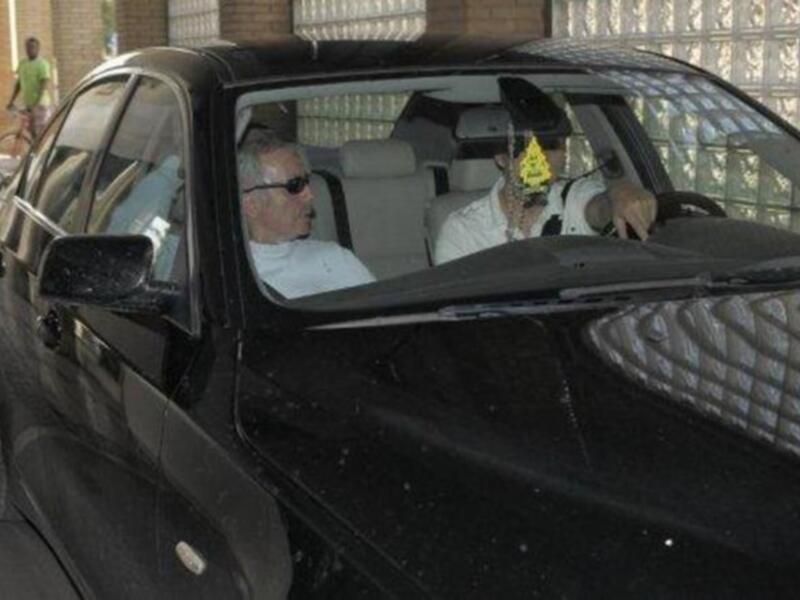 Un hombre con una gorra amarilla en el parabrisas de un coche negro, mientras otro hombre le toma una foto.
