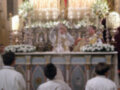 Un grupo de sacerdotes vestidos con hábitos blancos se encuentran en una iglesia, frente a un altar grande y elaborado. El altar está adornado con flores blancas y varias velas encendidas, creando una atmósfera de reverencia. En el fondo, se puede ver un altar menor con una imagen venerada y un manto rojo.