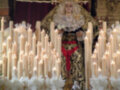 La Virgen del Rosario, vestida con un manto dorado y una corona de espinas, se encuentra en el centro de la imagen. A su alrededor, hay numerosas velas encendidas y flores blancas, creando un ambiente de reverencia y devoción. La escena se desarrolla en una iglesia con detalles arquitectónicos y un altar visible al fondo.
