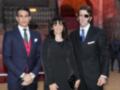Tres personas posan en un evento formal, dos hombres con trajes y una mujer con abrigo negro. La escena parece ser en un lugar histórico o ceremonial, con una arquitectura de fondo.