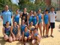 Equipo de natación femenina posando en la piscina, con uniformes azules y blancos, junto a dos entrenadores en el fondo.
