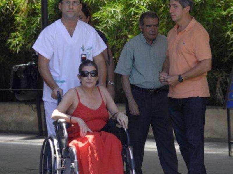 Una persona en silla de ruedas, vestida con un vestido rojo, es acompañada por tres hombres: uno en uniforme médico, otro en camisa verde y el tercero en polo naranja. La escena parece ser al aire libre, con vegetación y un edificio en el fondo.