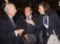 Tres personas en trajes, uno de ellos parece ser un político italiano. La imagen muestra una reunión formal con asistentes en el fondo.