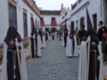 Un grupo de personas vestidas con trajes tradicionales andaluces caminan por una calle adoquinada. La imagen muestra detalles de la arquitectura local, con edificios blancos y rojos a ambos lados de la calle.