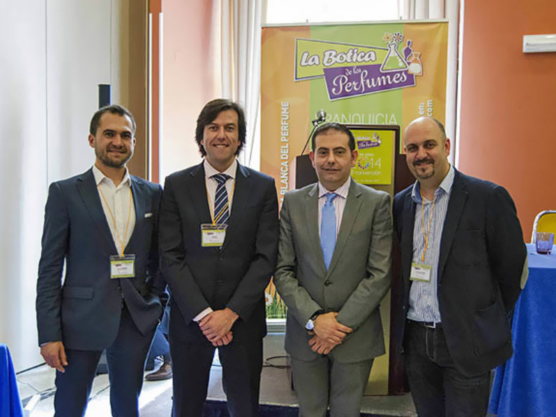 Tres hombres en trajes posan frente a un cartel de "La Botica" y "Perfumes", en una conferencia o evento profesional.