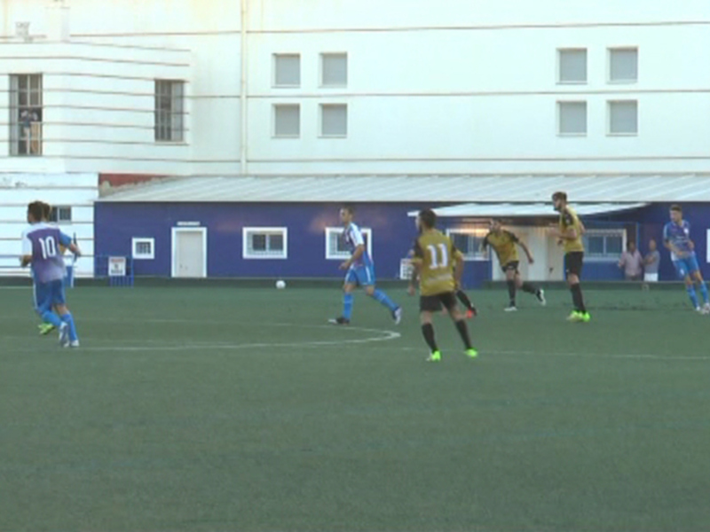 Un partido de fútbol en curso con jugadores en acción. El campo es verde y el estadio tiene una fachada blanca con ventanas. Los jugadores están usando uniformes de diferentes colores, uno en azul y otro en amarillo. La acción parece estar en el centro del campo, con un jugador de azul corriendo hacia la pelota.