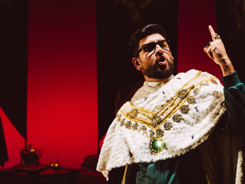 Un hombre con vestimenta tradicional y joyas de oro, posiblemente un personaje histórico o ceremonial, está en una escena teatral con fondo rojo.