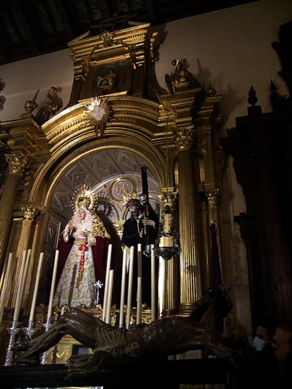 Altar de una iglesia con estatuas religiosas y un crucifijo en el centro, iluminado por una lámpara de araña.