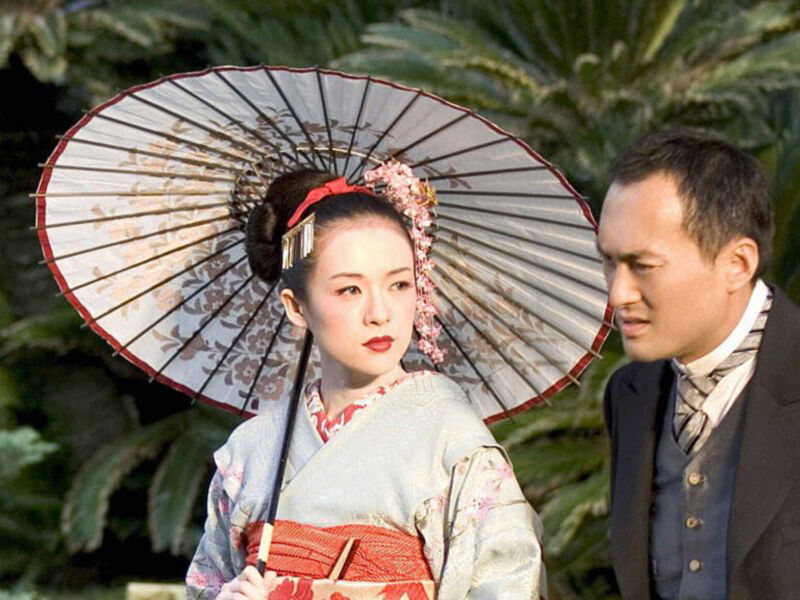 Imagina un par de personajes en medio de una escena histórica. La mujer, vestida con un kimono tradicional japonés y un sombrero de paja, mira hacia la derecha con expresión seria. El hombre, vestido con un traje formal, también mira hacia la derecha. Ambos están en un entorno natural con plantas verdes al fondo, creando una atmósfera de pasado.