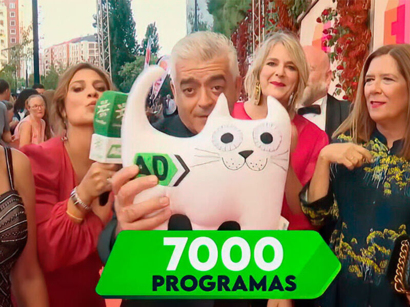 Una imagen de una celebración en un escenario con varias personas sonriendo y posando. Algunas están sosteniendo una figura de gato blanco con un mensaje verde que dice "7000 programas". La escena parece ser parte de una celebración o evento festivo.