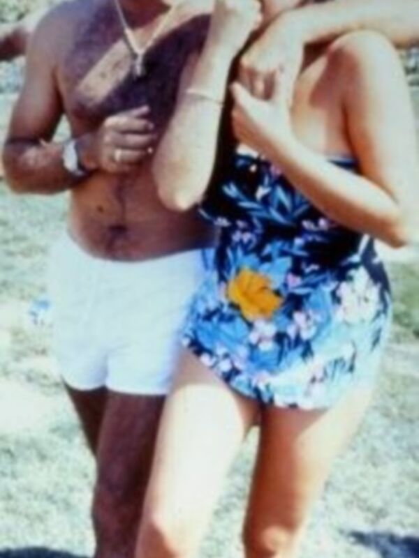 Un hombre y una mujer caminan juntos en la playa. La mujer lleva un traje de baño floral y el hombre una camiseta blanca con pantalones cortos. Ambos sonríen y parecen disfrutar de su tiempo libre en la playa.