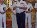 En una ceremonia naval, un oficial de uniforme blanco y negro con insignias reina el centro. Detrás, una bandera española ondea en un altar dorado con detalles artísticos. La escena transmite solemnidad y formalidad, probablemente en un evento oficial de la marina.