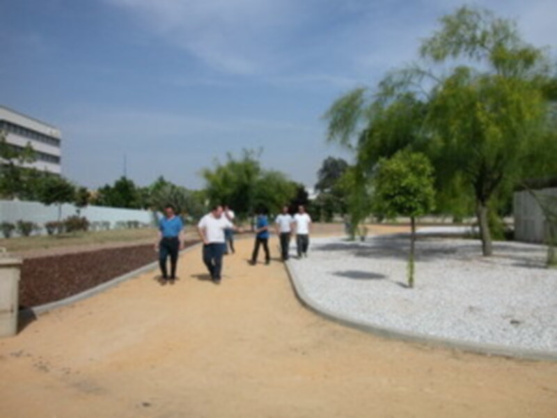 Un grupo de personas caminan por un camino de tierra en un entorno verde con árboles y vegetación. El cielo es azul claro, indicando buen tiempo. La imagen parece ser de un campus o parque público.