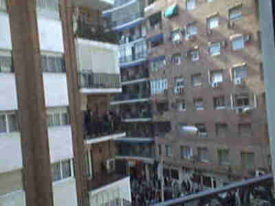 Imagen de un edificio con varios pisos y balcones. La perspectiva es desde una ventana, mostrando la urbanización y la vida cotidiana de las personas en el barrio.