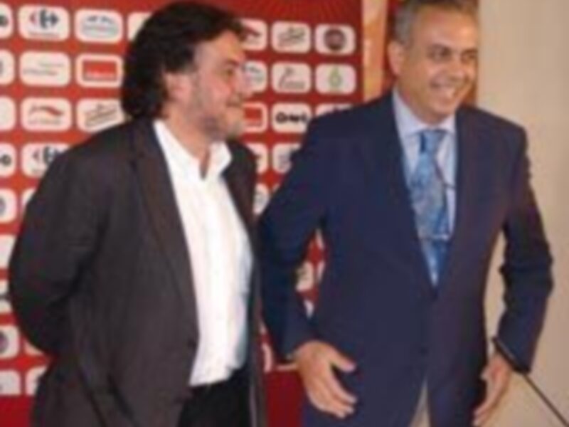 Dos hombres posan frente a un fondo rojo con un logo en el centro, uno de ellos con una camisa blanca y el otro con una camisa azul y corbata azul claro.
