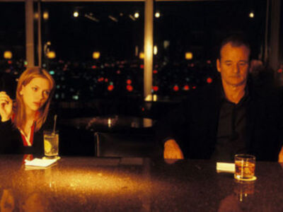 Una mujer y un hombre están sentados en una mesa de cristal, disfrutando de bebidas alcohólicas. La imagen está iluminada con luces brillantes en el fondo, creando un ambiente romántico.