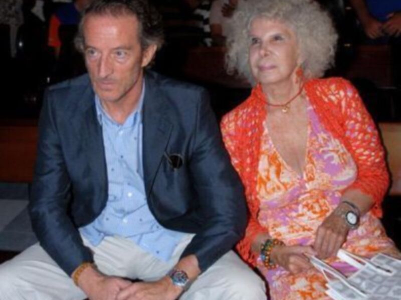 Una pareja sentada, el hombre con una camisa azul y el hombre con una camisa azul y pantalones blancos, la mujer con un vestido naranja y una corona de flores, ambos sonriendo y mirando hacia la cámara.