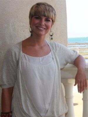 A una mujer con cabello corto y rubio, llevando un vestido blanco suelto. Está sonriendo y posa en una estructura blanca con un fondo que parece ser la playa.
