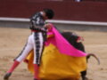 Un torero se enfrenta a un toro en una plaza de toros, usando un capote y un chaleco negro con detalles plateados. La escena se desarrolla en una arena de lucha, con el suelo cubierto de tierra y la pared del ruedo visible en el fondo.