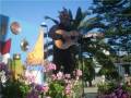 Un hombre toca la guitarra en un parque con flores y palmeras en el fondo.