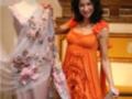 Una mujer posa con dos vestidos: uno de encaje rosa con flores y un otro de color naranja. El fondo es una sala con muebles clásicos.