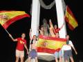 Jóvenes celebrando con banderas españolas frente a una escultura monumental en la noche.