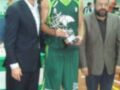 Un hombre en camiseta verde y pantalones cortos, con un trofeo en la mano, posa junto a otros dos hombres. El fondo muestra una cancha de baloncesto con anuncios y un cartel que dice "Miguel".