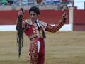 Un matador de toros en el ring, con traje tradicional rojo y dorado, sostiene un capote y una espada. El fondo muestra la arena de toros con barda roja y espectadores en las gradas.