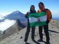 Dos personas en el pico de un volcán, sosteniendo una bandera verde con un diseño blanco y negro. El paisaje es desértico, con rocas y nieve en el fondo.