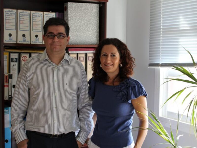 Dos personas posan en un entorno profesional con estanterías de archivos y una ventana con persianas.