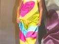 La modelo presenta un vestido de colores vibrantes, con una combinación única de amarillo, rosa y azul. El diseño es moderno y creativo, destacando la moda contemporánea en un evento de desfile.