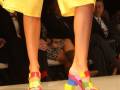 Las piernas de una modelo lucen un vestido amarillo brillante y sandalias coloridas con bloques de colores en el tacón, en un escenario de desfile.