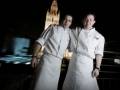 Dos chefs posan frente a la catedral de Westminster en Londres, iluminada por la noche.
