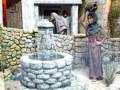 Figura histórica en escena medieval, con mujer levantando jarra cerca de pozo.