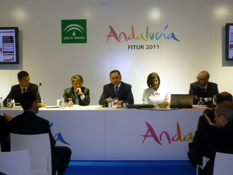 Una conferencia en Andalucía FITUR 2011, con cuatro personas sentadas detrás de una mesa blanca. La pared tiene un logotipo verde y letras coloridas que dicen "Andalucía FITUR 2011".