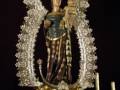 Estatua de una Virgen con un niño en sus brazos, rodeada por flores y adornos dorados.
