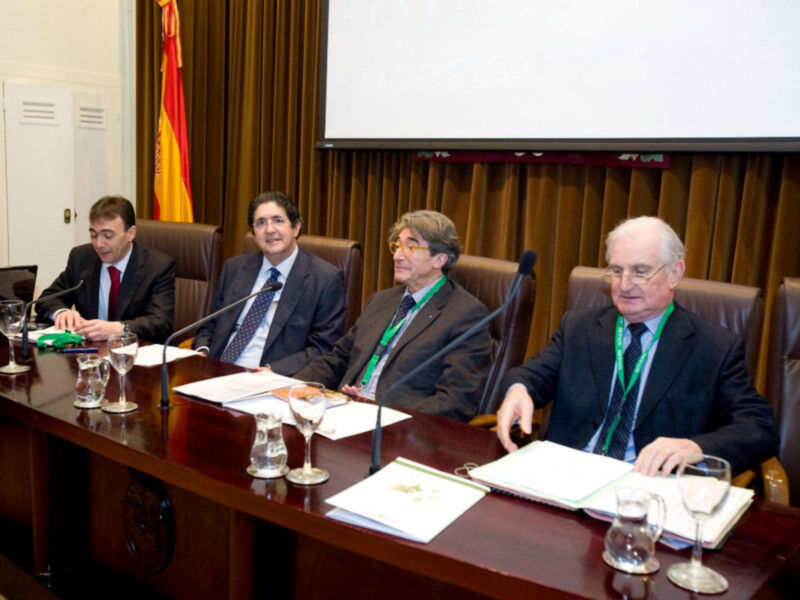 Conferencia o reunión formal en un salón con bandera española, hombres en trajes sentados en un escritorio con documentos y micrófonos.