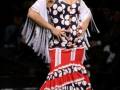 Una modelo desfilando con un vestido de flamenco en blanco, rojo y negro con lunares blancos. La falda es larga y cóncava, con una capa blanca con flecos. La modelo lleva un tocado de flores rojas y una corona en la cabeza.