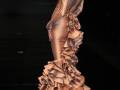 Modela en desfile con vestido dorado y falda volante, complementado con sombrero y pendientes grandes.