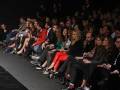 Una audiencia en un evento de moda, sentada en filas alineadas. La mayoría están vestidos formalmente y observan atentamente la pasarela. El ambiente es oscuro, destacando los colores brillantes de las prendas y la atención de los asistentes.