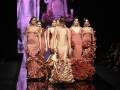 Una modelo con vestido de flamenca camina por la pasarela, seguida de otras modelos en vestidos similares. La escena está iluminada con una pantalla LED en el fondo que muestra imágenes de flores. La imagen captura la atmósfera de un desfile de moda, con el público observando atentamente.
