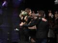 Una mujer en un vestido negro con mangas transparentes y detalles de encaje, abraza a una persona masculina también vestida con negro. Ambos están en un escenario iluminado, con una pantalla de fondo que muestra imágenes de luces. La escena sugiere un evento formal o celebración, posiblemente un concierto o una gala.