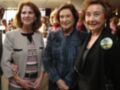 Tres mujeres sonrientes posan juntas en un evento. La imagen muestra una celebración o reunión formal con vestimenta elegante y una insignia en la chaqueta de la mujer a la derecha.