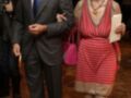 Un hombre y una mujer caminan juntos en un evento formal, el hombre viste un traje y la mujer lleva un vestido de rayas rojas y beige.