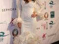 Eva Longoria posa en el escenario de la moda internacional en una elegante vestidura blanca con detalles florales y mangas largas. En el fondo, se pueden ver logos de diferentes marcas y eventos.